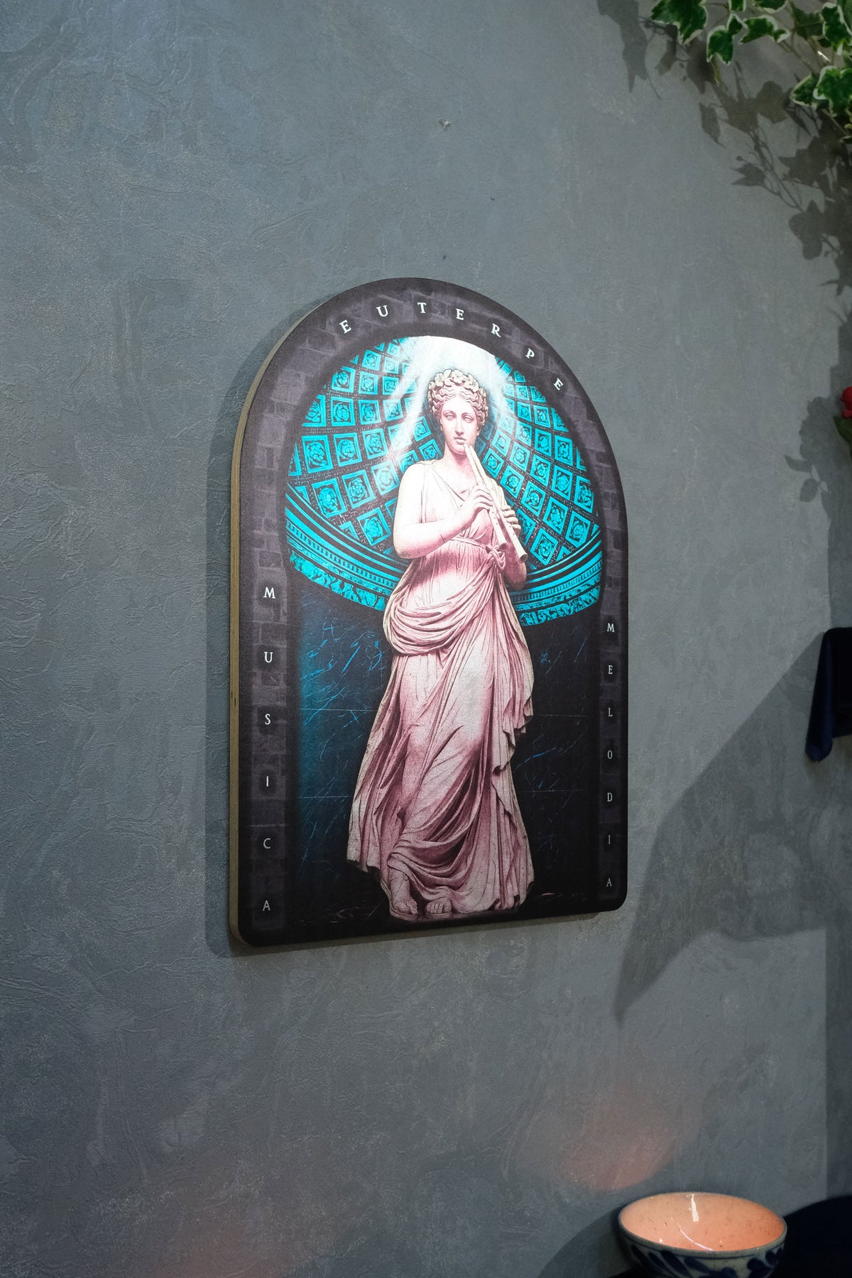 An arched decorative wall art piece featuring a classical female figure in flowing pale pink drapery against a glowing teal and black geometric background, mounted on a textured gray wall. Euterpe — Muse of Music