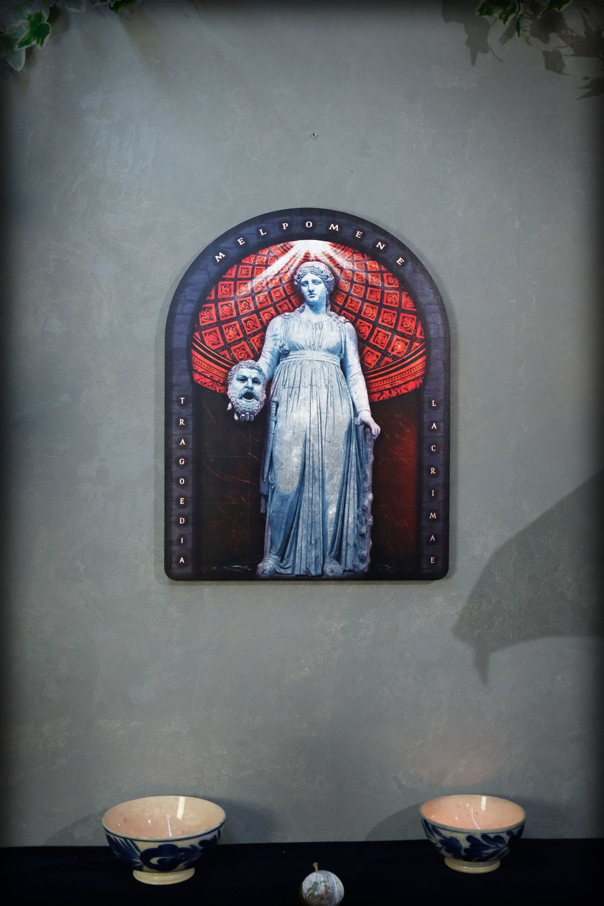 A framed gothic-style wall art piece featuring a pale blue statue of a woman holding a skull, set against a red patterned arch, mounted on a textured gray wall above a dark surface with two small bowls.dark academia decor Melpomene
Muse of Tragedy