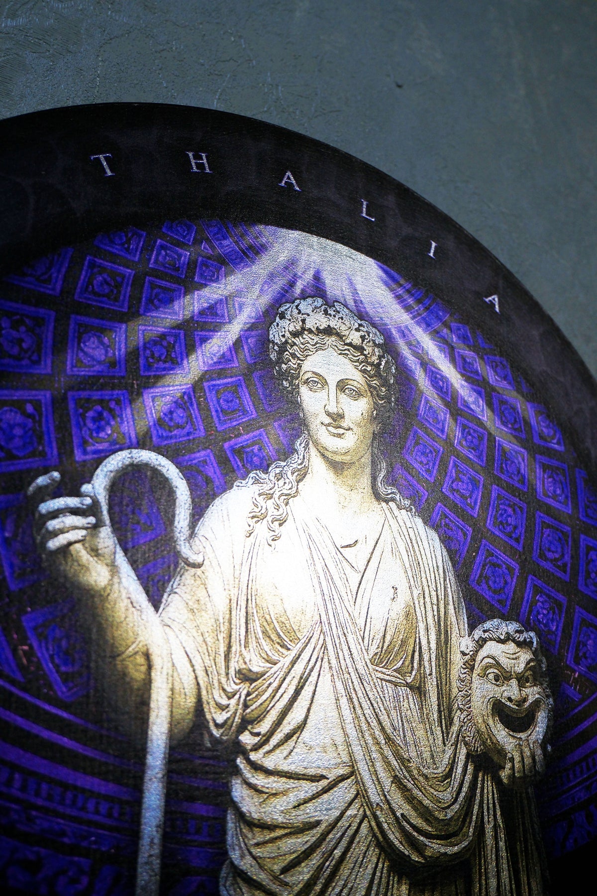 A close-up of a circular stained glass window featuring a classical statue of Thalia, the goddess of speech, holding a snake and a lion head. The background of the design is a deep blue and purple patterned grid, with the name 'THALIA' arched above in white lettering. Thalia. Muse of Comedy