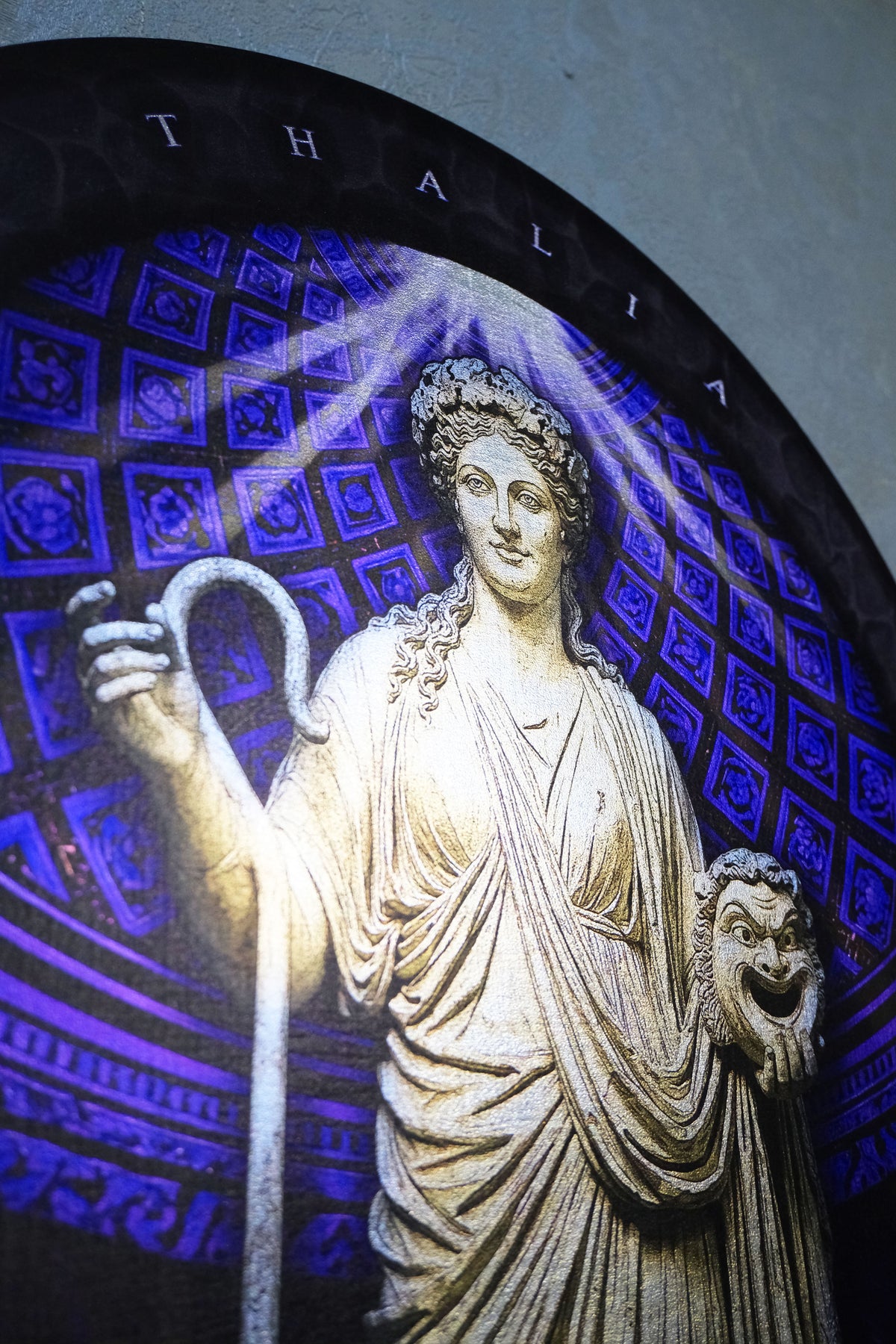A close-up of a stained glass window depicting a classical female figure in cream drapery holding a staff, set against a blue and purple patterned background with the name THALIA at the top. Thalia. Muse of Comedy