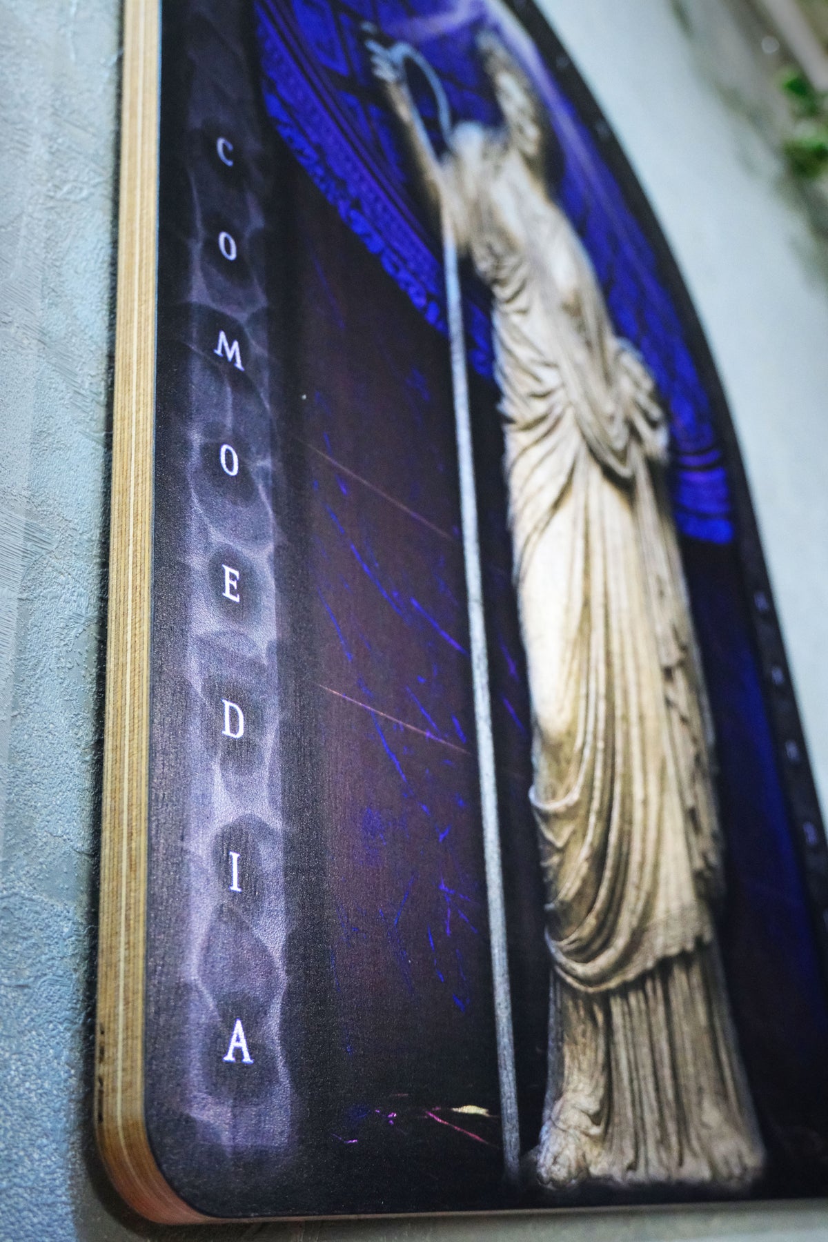 A close-up angled shot of a decorative sign or plaque featuring the word 'COMEDIA' in white letters over a vertical X-ray style spine, next to a classical statue in beige tones against a dark blue and black background. Thalia. Muse of Comedy