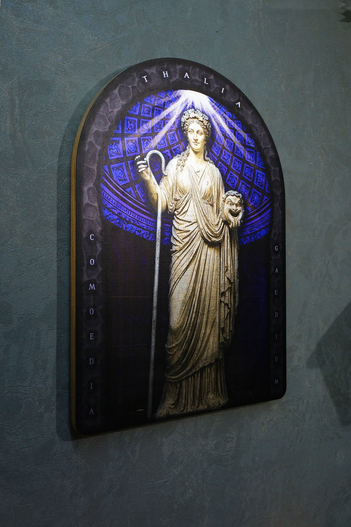 An illuminated decorative wall plaque featuring a classical statue of Thalia in a gold-toned relief, set against a glowing blue patterned background with a dark arched frame, mounted on a textured gray wall. Thalia. Muse of Comedy