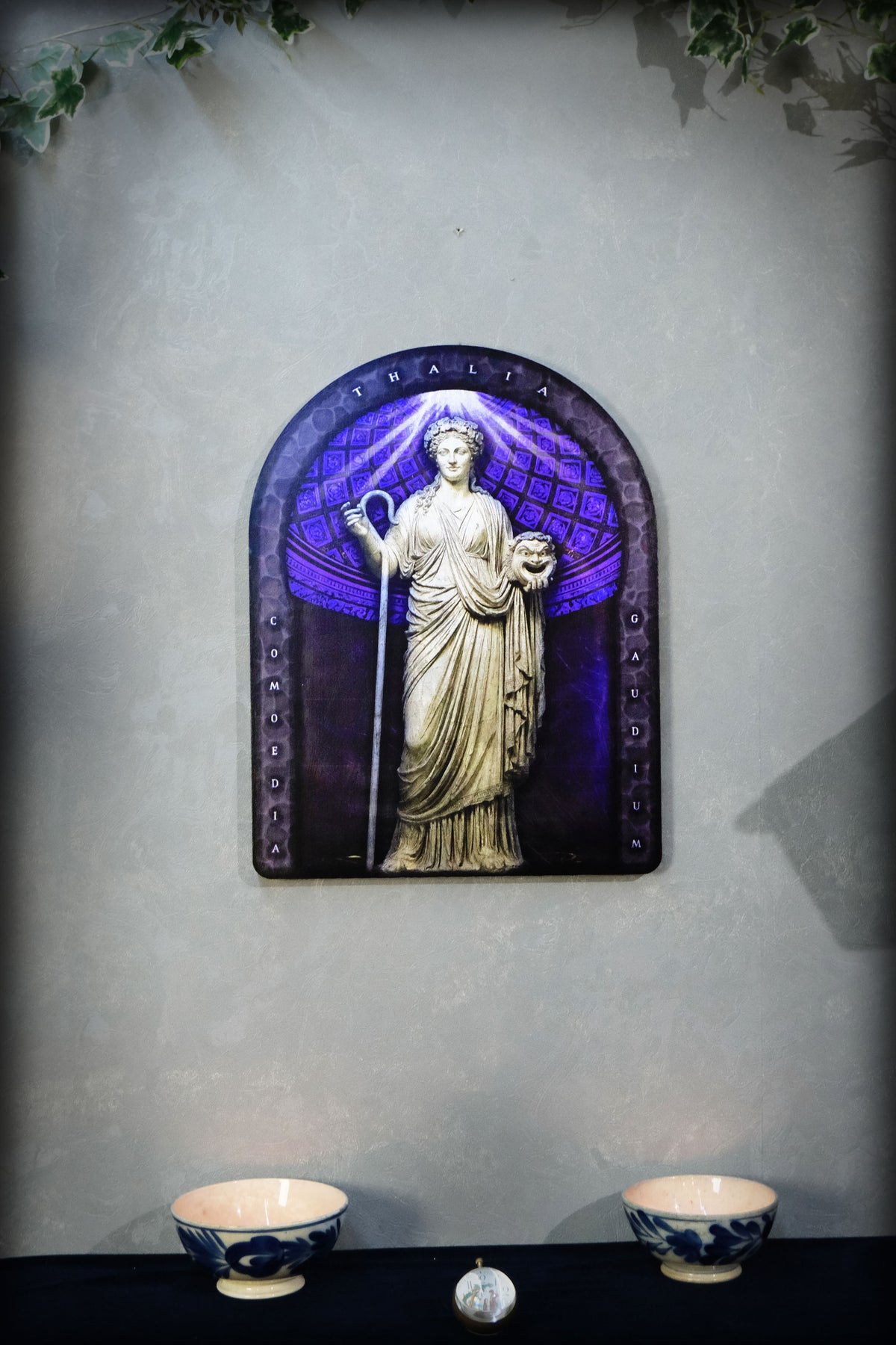 A decorative wall plaque featuring a classical statue of a robed female figure holding a staff, set against a purple and blue arched background. The plaque is mounted on a textured gray wall above a black surface with two small blue-and-white bowls. Thalia. Muse of Comedy