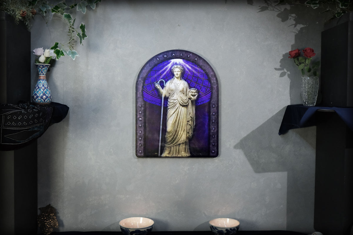 A decorative wall-mounted arched plaque featuring a classical statue in gold tones set against a purple and blue stained-glass-style background, displayed on a textured gray wall with shelves and flowers flanking it. Thalia. Muse of Comedy