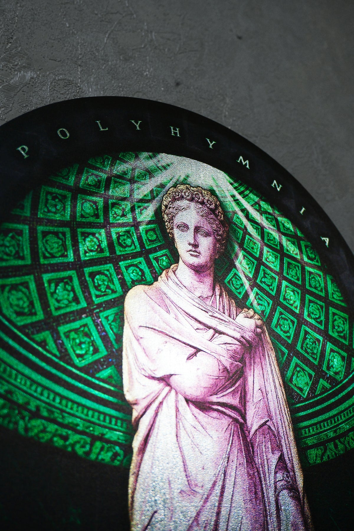 A circular decorative plaque or print featuring a classical statue of a draped female figure in pinkish-white tones, set against a green patterned background with the word 'POLYHYMNI'A at the top, photographed against a dark textured wall. Polyminia is a common alternative spelling of Polyhymnia (or Polymnia), the Greek Muse of sacred poetry, hymns