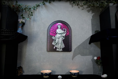 A framed wall art piece featuring a white classical statue figure against a pink and purple arched background, mounted on a textured gray wall above a dark table with two lit candles. dark academia decor  Terpsichore — Muse of Dance