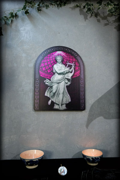 An arched decorative wall plaque featuring a classical statue in white against a pink and purple patterned background, mounted on a textured gray wall above a dark surface with two lit bowls and a small round object. dark academia decor  Terpsichore — Muse of Dance