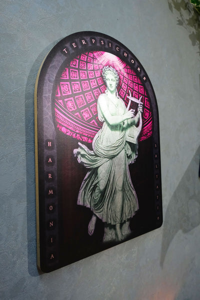 A decorative arched wall plaque featuring a classical female figure in flowing drapery holding a book, set against a pink geometric pattern on a dark background. The plaque is mounted on a textured light gray wall. dark academia decor  Terpsichore — Muse of Dance