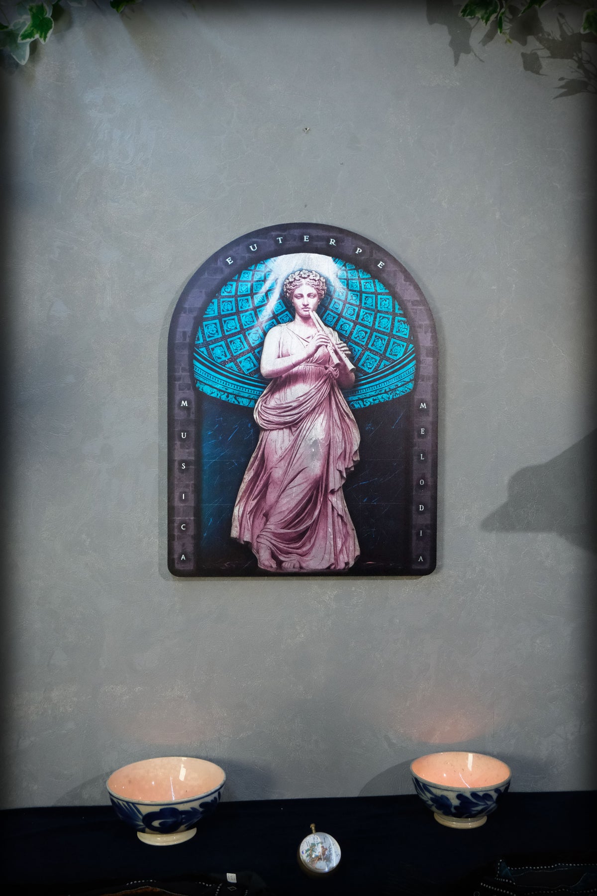 An arched decorative wall art piece featuring a classical female figure in pink drapery against a teal globe-like background, mounted on a textured gray wall above a dark table with two blue-and-white bowls. Euterpe — Muse of Music