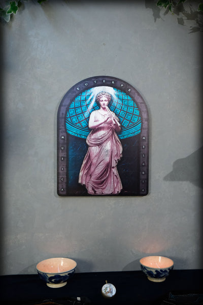 An arched decorative wall art piece featuring a classical female figure in pink drapery against a teal globe-like background, mounted on a textured gray wall above a dark table with two blue-and-white bowls. Euterpe — Muse of Music