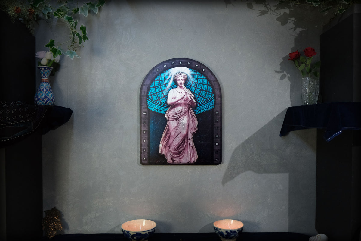 A framed wall art piece featuring a classical statue of a woman in pink drapery against a blue circular background, mounted on a textured gray wall above two lit candles, with decorative vases and greenery in the background. dark academia decor  Euterpe — Muse of Music