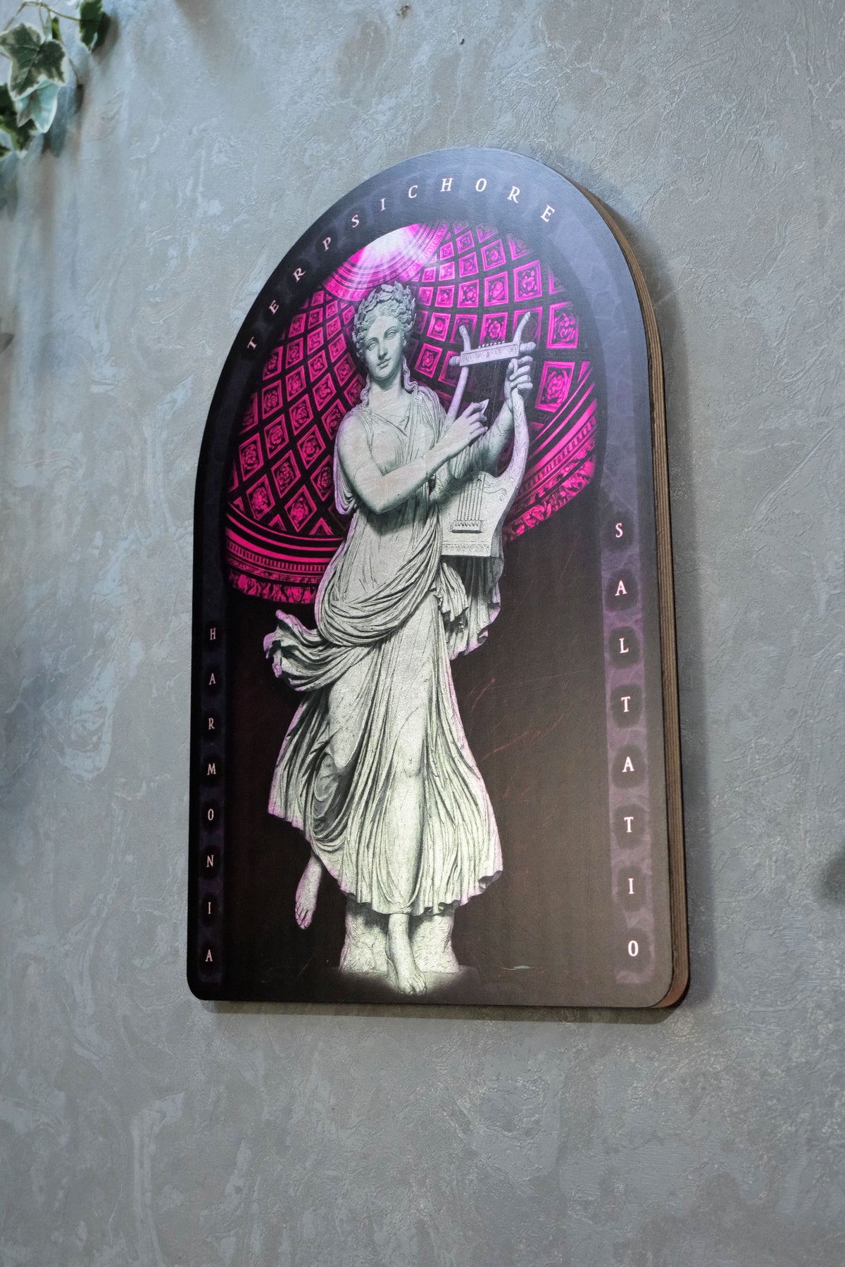An arched decorative wall plaque featuring a classical statue illustration in white and gray against a pink and black patterned background, mounted on a textured gray wall. dark academia decor  Terpsichore — Muse of Dance