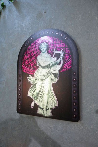 An arched decorative plaque featuring a classical statue of a draped female figure holding a lyre, set against a pink geometric pattern on a dark background, mounted on a textured gray wall. dark academia decor  Terpsichore — Muse of Dance