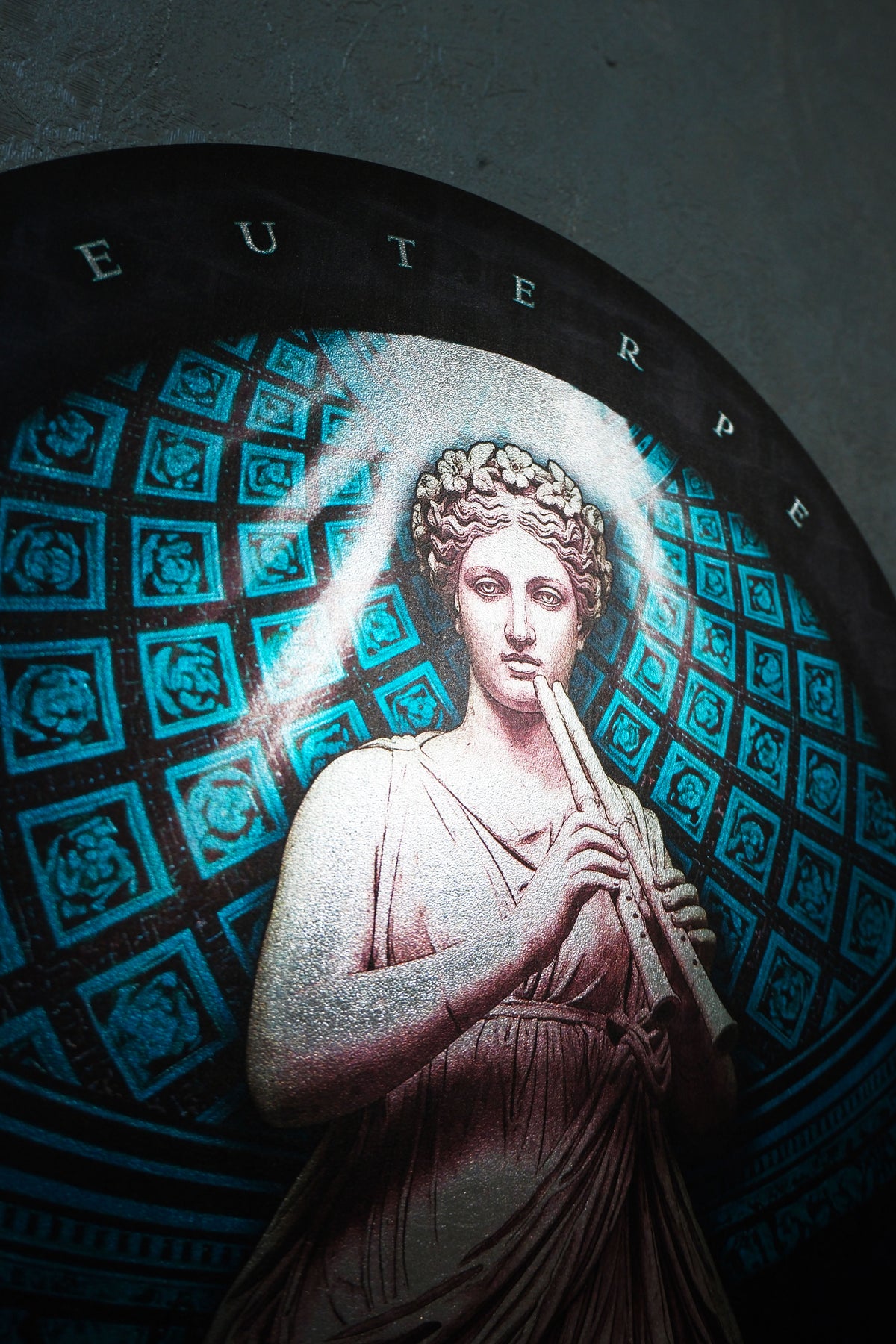 A circular stained glass window depicting the classical figure of Euterpe, holding a flute, with a teal geometric patterned background and a dark arched frame. Euterpe — Muse of Music