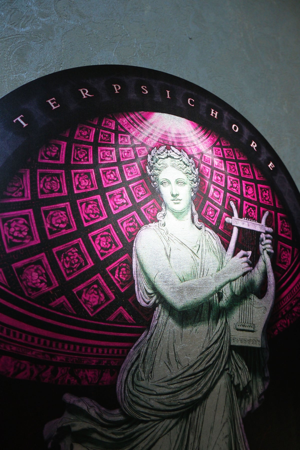 A circular decorative plaque or wall art featuring a classical female figure in a flowing robe holding a lyre, set against a pink and black geometric pattern. The word 'TERPSICHORE' appears along the top edge. dark academia decor  Terpsichore — Muse of Dance