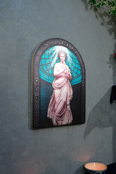 An arched decorative wall art piece featuring a classical female figure in flowing pale pink drapery against a glowing teal and black geometric background, mounted on a textured gray wall. Euterpe — Muse of Music