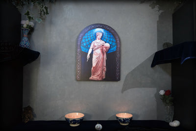 A framed religious wall art piece depicting the Virgin Mary in a pink robe and blue halo, mounted on a textured gray wall above a dark table with two lit bowls and small decorative objects. Urania is the Greek Muse of astronomy and astrology