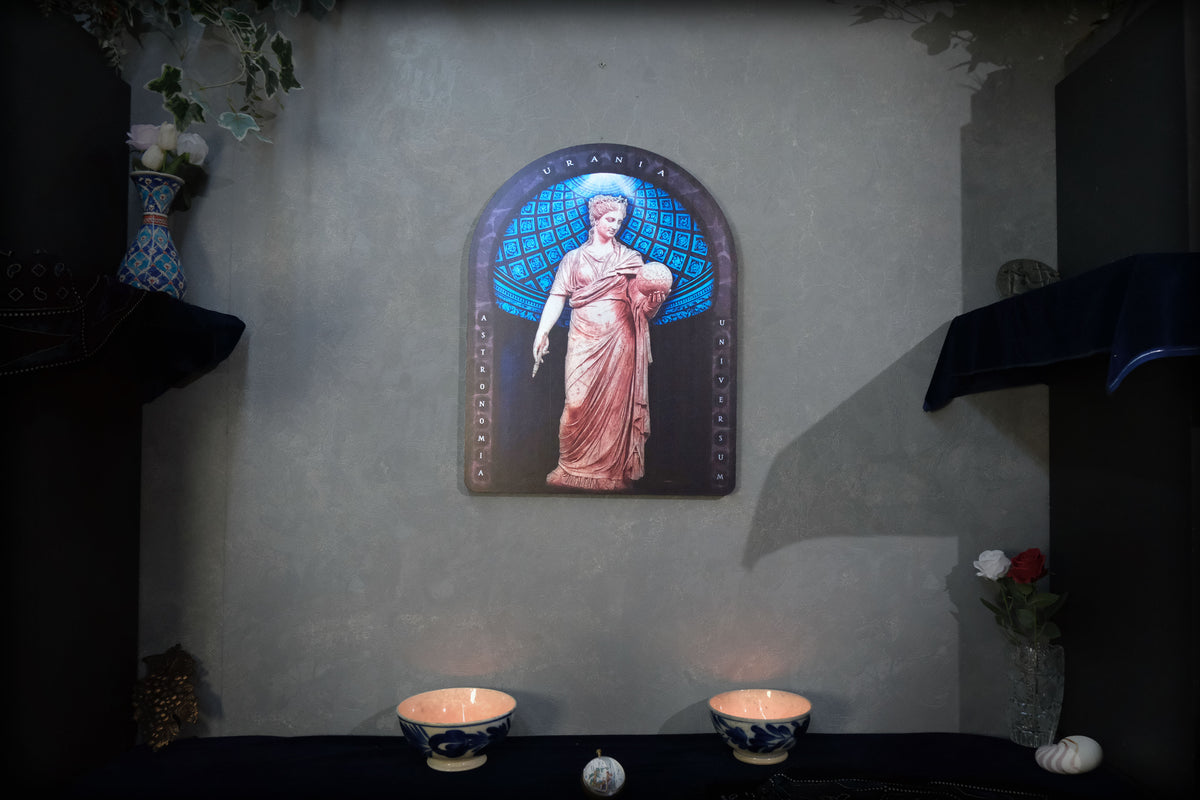 A framed religious wall art piece depicting the Virgin Mary in a pink robe and blue halo, mounted on a textured gray wall above a dark table with two lit bowls and small decorative objects. Urania is the Greek Muse of astronomy and astrology