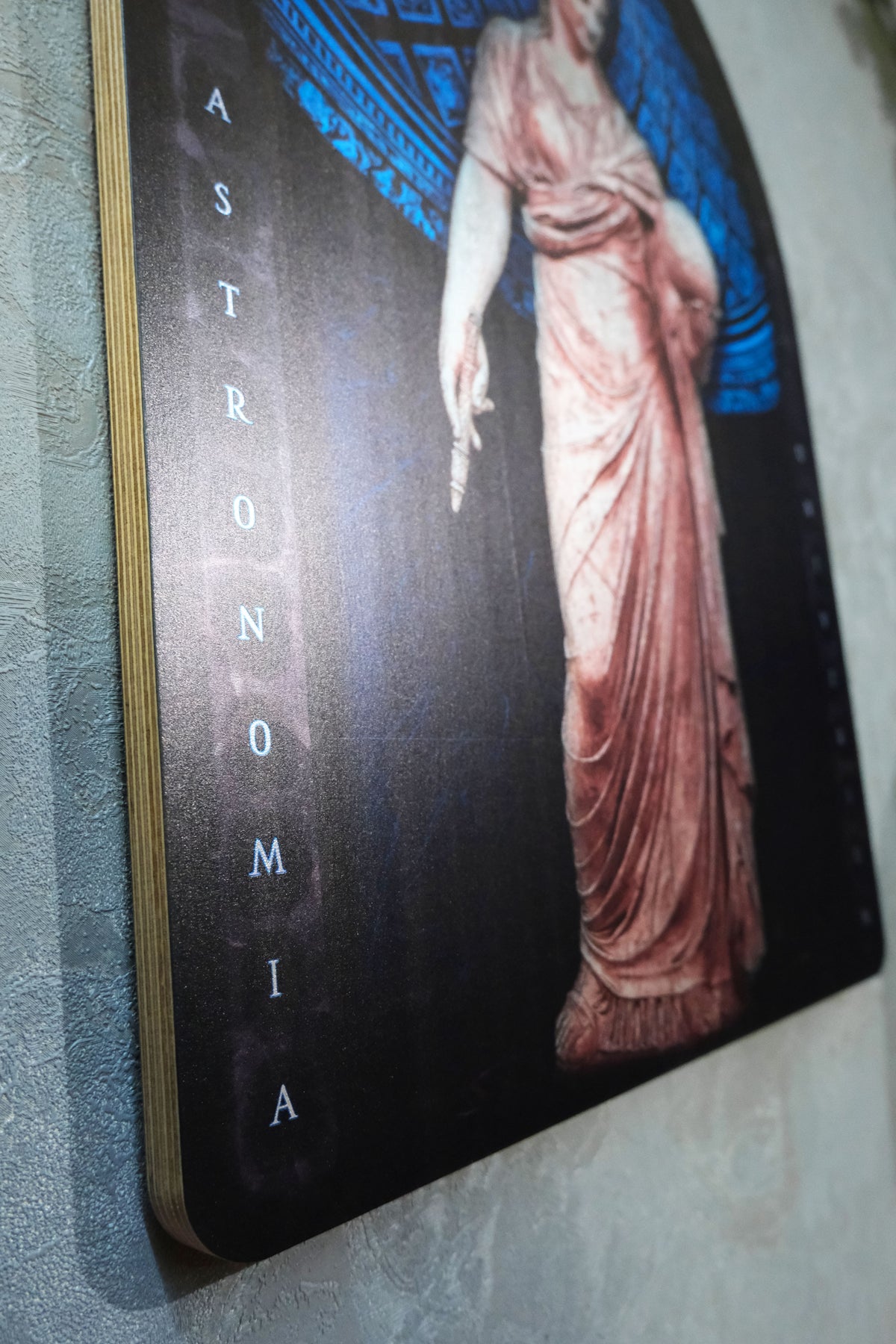 A close-up of a wall-mounted art print or poster featuring a classical draped figure in pinkish-beige tones against a dark background with blue decorative elements and the word 'ASTRONOMIA' printed vertically. Urania is the Greek Muse of astronomy and astrology
