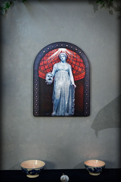A framed gothic-style wall art piece featuring a pale blue statue of a woman holding a skull, set against a red patterned arch, mounted on a textured gray wall above a dark surface with two small bowls.dark academia decor  Melpomene
Muse of Tragedy