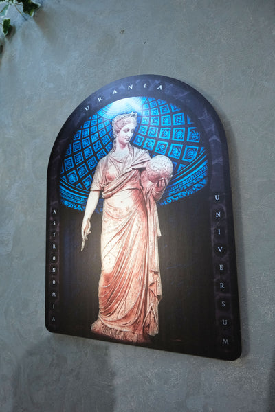 A decorative arched plaque or sign featuring a classical statue illustration with blue geometric patterns in the background and the words 'URANIA' at the top and 'UNIVERSUM' on the side, mounted against a textured gray wall. Urania is the Greek Muse of astronomy and astrology