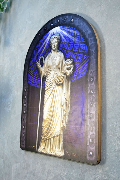 An arched decorative plaque featuring a classical statue of a draped female figure holding a staff and a lion, set against a glowing blue patterned background. The plaque has a dark border with the words 'THALIA', 'GABRIELLA', and 'GAUDIIUM' and is mounted on a textured gray wall. Thalia. Muse of Comedy