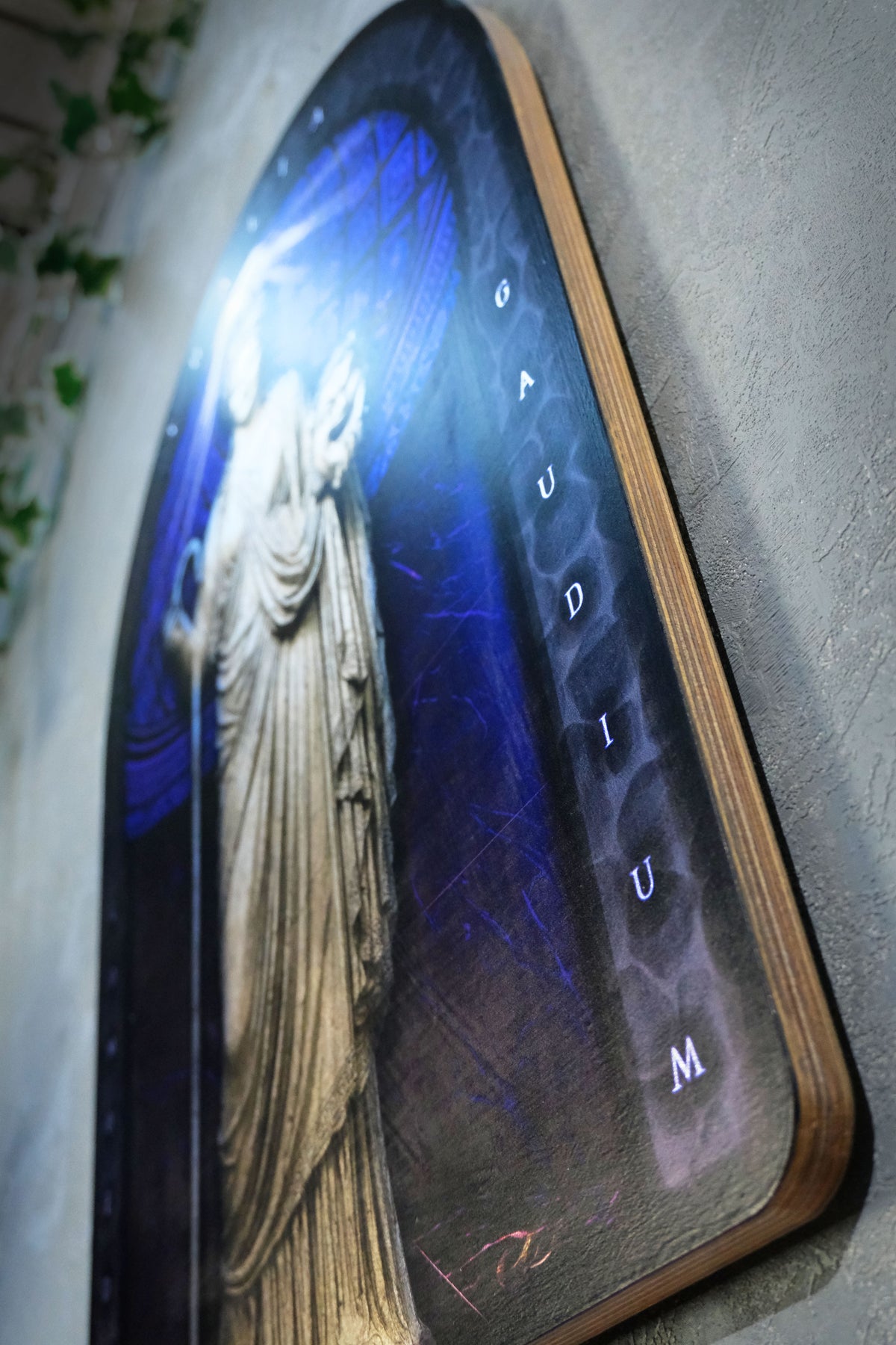 An arched wooden plaque featuring a glowing classical statue on a blue background with the word 'Gaudium' spelled out vertically in letters, mounted on a textured gray wall. Thalia. Muse of Comedy