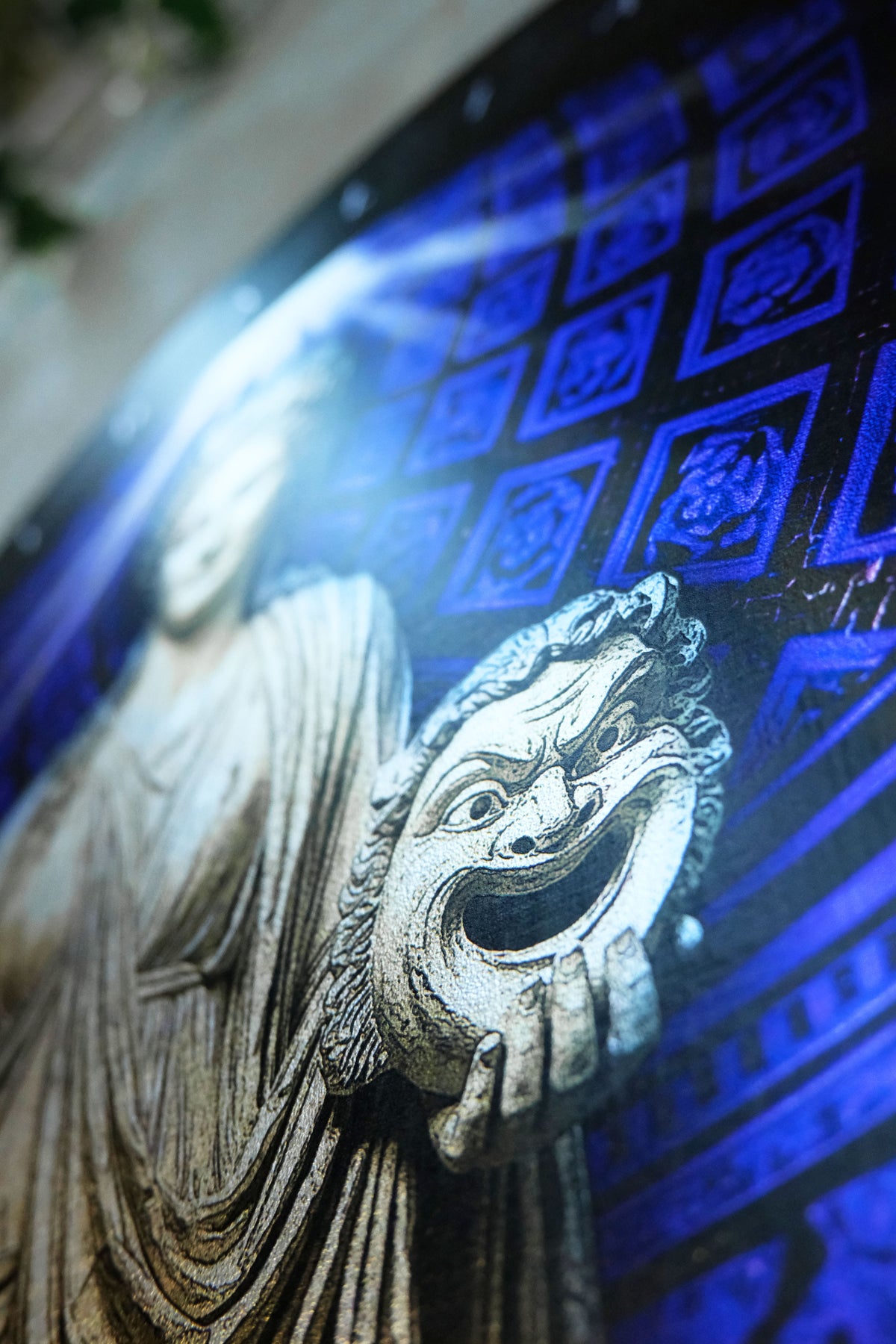 A close-up of a decorative wall art piece featuring a carved stone-like figure in gray holding a theatrical comedy and tragedy mask, set against a vivid blue patterned background. Thalia. Muse of Comedy