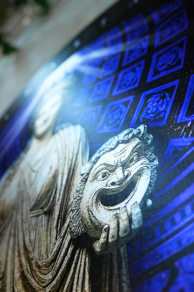 A close-up of a decorative wall art piece featuring a carved stone-like figure in gray holding a theatrical comedy and tragedy mask, set against a vivid blue patterned background. Thalia. Muse of Comedy