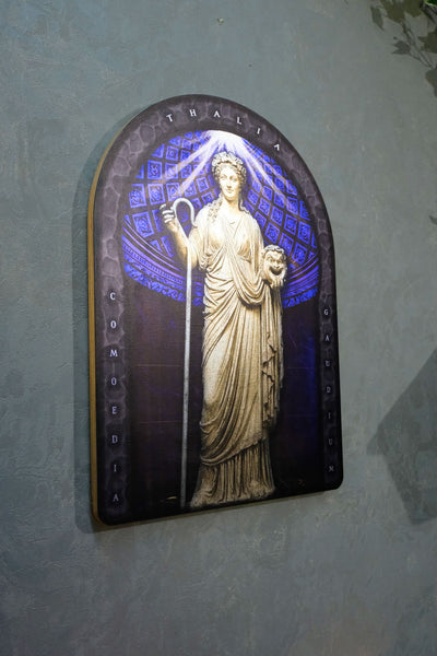An arched decorative plaque featuring a classical statue of a woman in a robe holding a staff, set against a blue patterned background with the words 'THE ALIA' and 'COMEDIA' on the sides, mounted on a textured gray wall. Thalia. Muse of Comedy