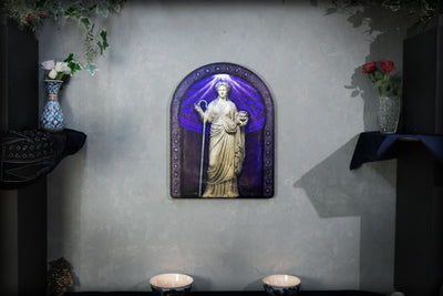 A decorative wall-mounted arched plaque featuring a classical statue in gold tones set against a purple and blue stained-glass-style background, displayed on a textured gray wall with shelves and flowers flanking it. Thalia. Muse of Comedy