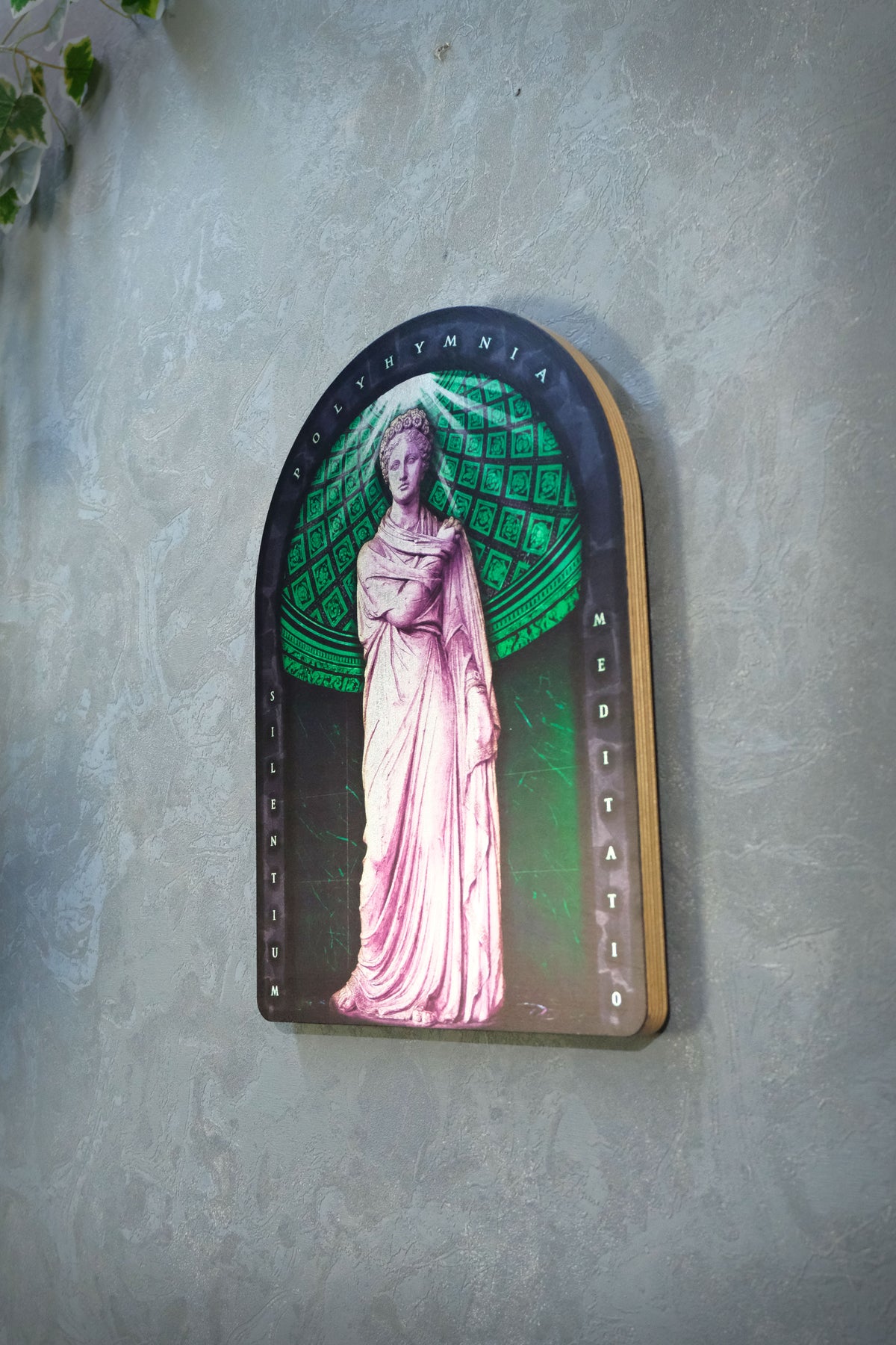 An arched decorative plaque featuring a classical statue in a pink gown against a green patterned background, mounted on a textured gray wall. Polyminia is a common alternative spelling of Polyhymnia (or Polymnia), the Greek Muse of sacred poetry, hymns