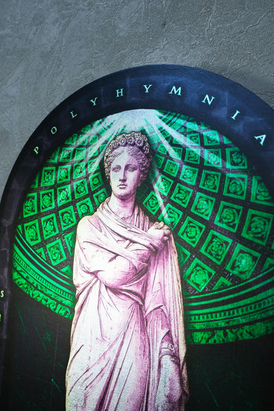 A circular wall art piece featuring a classical statue of a draped female figure in pinkish-white tones, set against a green geometric pattern and a dark arched top with the word 'POLYHYMnia' in white letters. Polyminia is a common alternative spelling of Polyhymnia (or Polymnia), the Greek Muse of sacred poetry, hymns