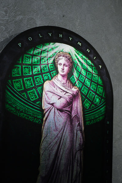 A stained glass window depicting a classical female figure in a draped gown, set against a green geometric pattern and framed by a black arch with the word 'POLYHYMNIA' at the top. Polyminia is a common alternative spelling of Polyhymnia (or Polymnia), the Greek Muse of sacred poetry, hymns