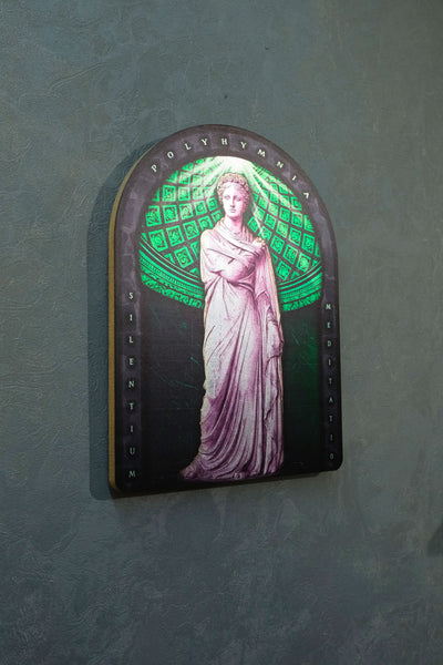 A gothic-style arched wall plaque featuring a statue of a woman in a flowing gown against a green patterned background, mounted on a textured gray wall. Polyminia is a common alternative spelling of Polyhymnia (or Polymnia), the Greek Muse of sacred poetry, hymns