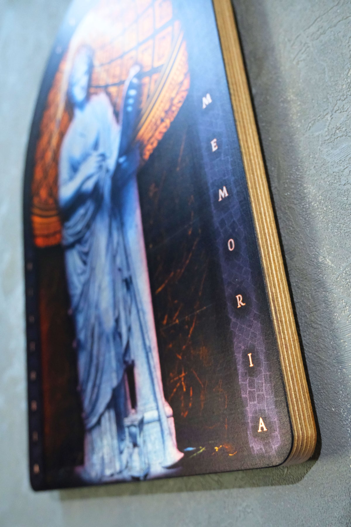 A close-up of a printed wooden plaque or book-like memorial item with a dark gothic illustration of a statue and the word 'MEMORIA' running vertically, photographed against a gray textured wall. dark academia decor  Clio — Muse of History