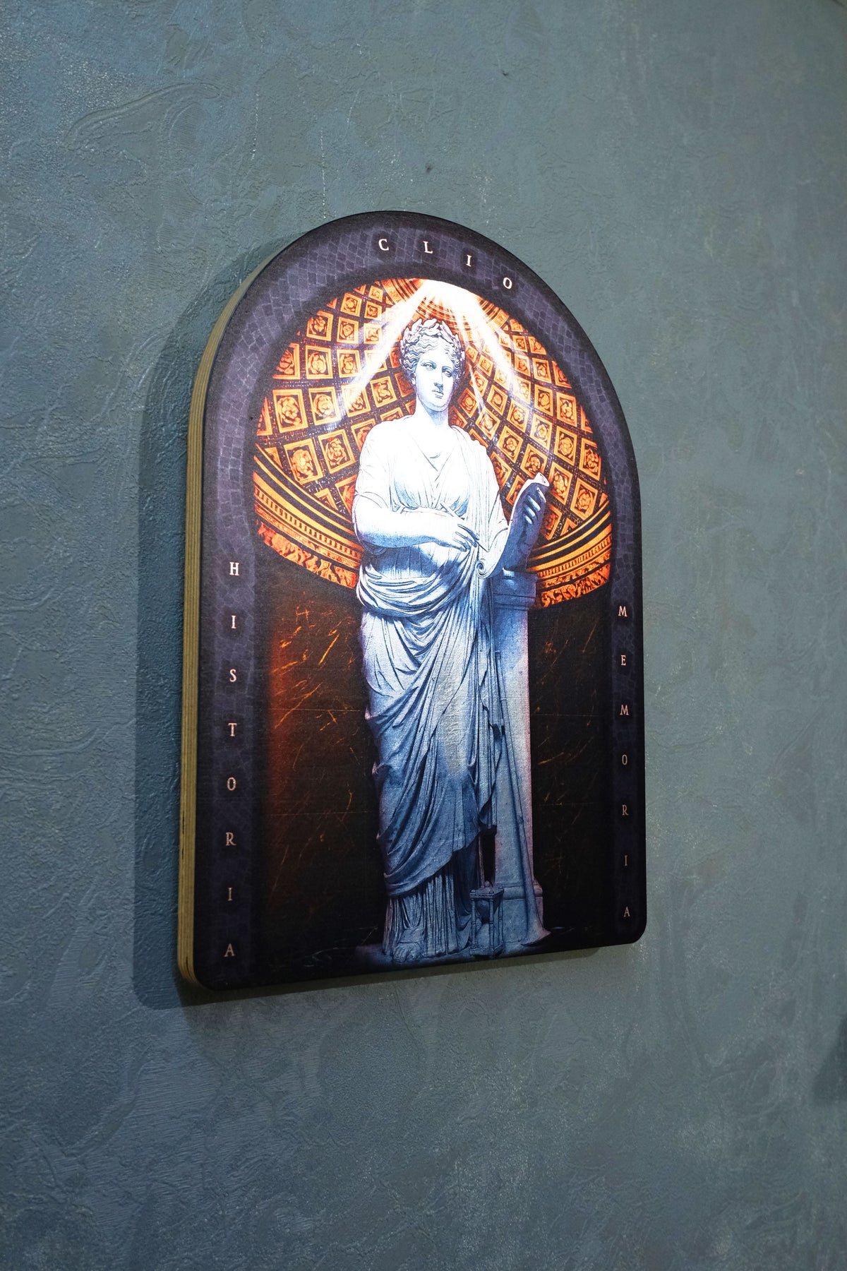 An arched decorative wall plaque featuring a classical white marble statue of a woman against an orange and gold patterned background, mounted on a textured blue-gray wall. dark academia decor  Clio — Muse of History