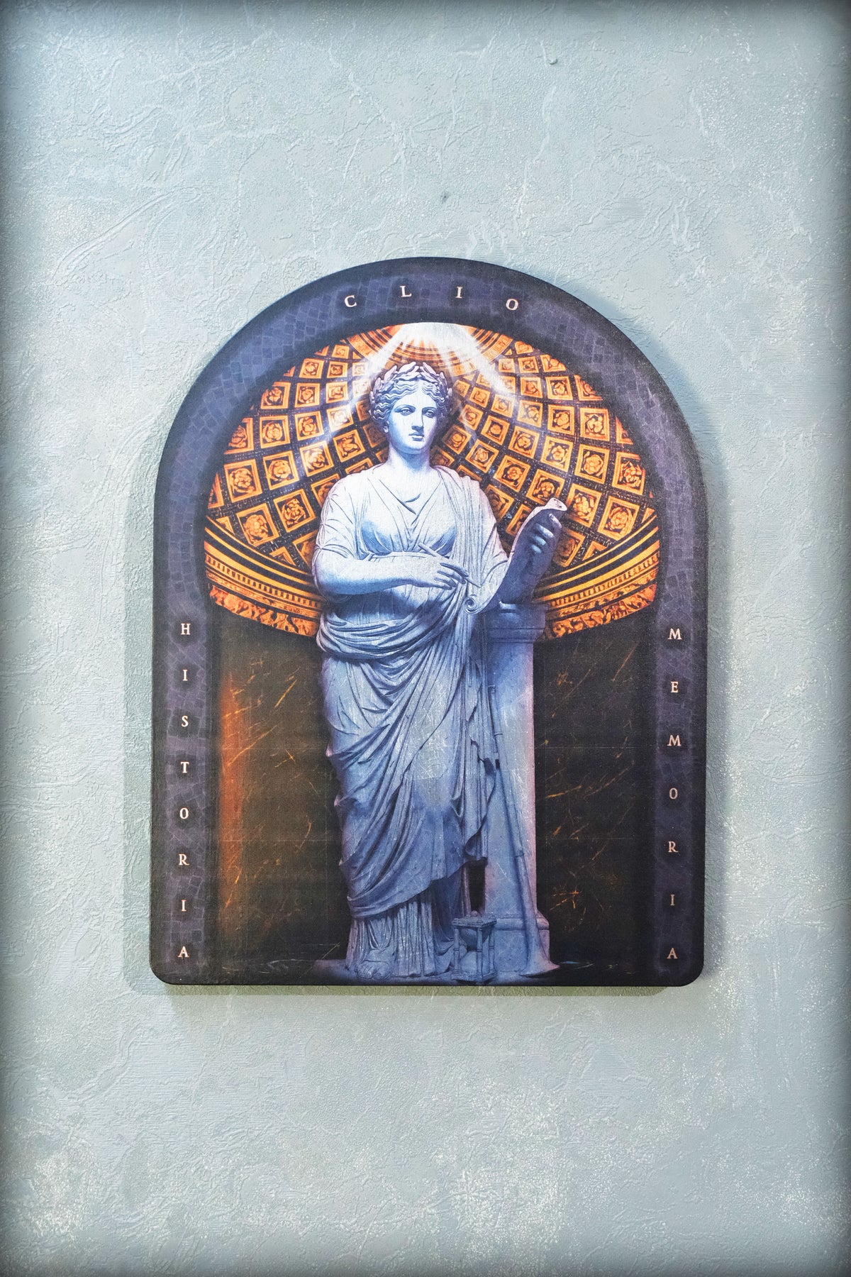 An arched decorative plaque featuring a classical statue of a draped female figure in blue-gray tones, set against a patterned gold and dark background, mounted on a textured light gray wall. dark academia decor  Clio — Muse of History