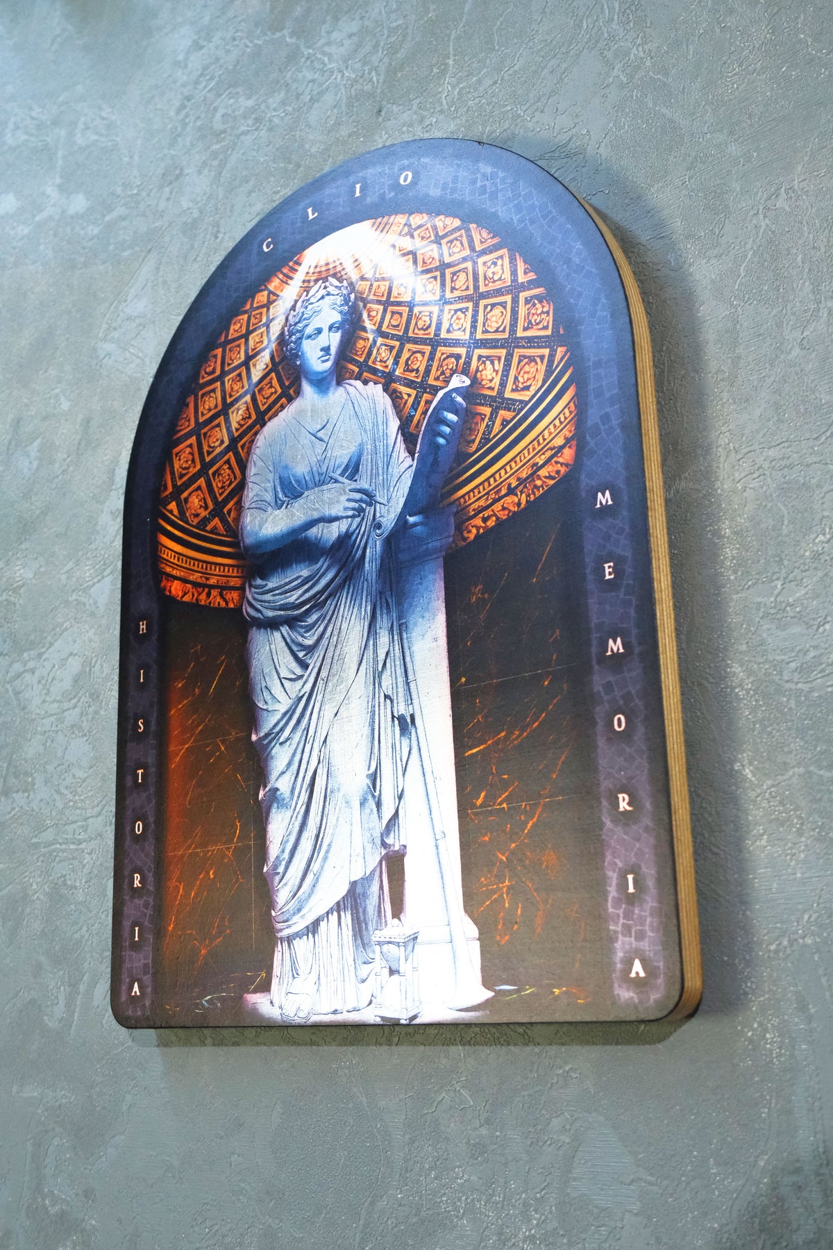 An arched decorative plaque featuring a blue-toned statue of a robed figure holding a book, with orange geometric patterns in the background and vertical text reading 'historia' on the left and 'MEMORIA' on the right, mounted against a textured gray wall. dark academia decor  Clio — Muse of History